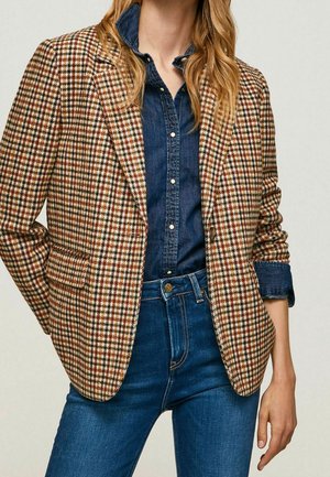 Femme portant un blazer à motif pied-de-poule marron et blanc, par-dessus une chemise en denim bleu foncé boutonnée, rentrée dans un jean en denim bleu.