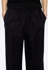 Schwarze, taillierte Hosen mit elastischem Bund, seitlichen Taschen und einer glatten, matten Oberfläche. Mit dezenten Nahtdetails und einer lockeren Passform.