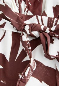 Tissu à motifs marron et blanc avec de grands motifs de feuilles, présentant un détail noué au centre et une surface légèrement texturée.