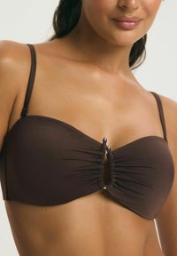 Kobieta w ciemnobrązowym bikini bandeau z cienkimi ramiączkami i centralną srebrną klamrą, pokazana od ramion do połowy tułowia.