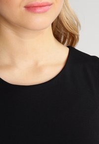 Närbild av en kvinnas nedre ansikte och hals med ljus hud, blont hår, iklädd en svart tröja med rund hals.