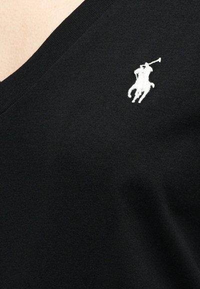 Κοντινό πλάνο μαύρου υφάσματος με λευκό κεντημένο λογότυπο παίκτη πόλο πάνω σε άλογο κοντά σε λαιμόκοψη σε σχήμα V.