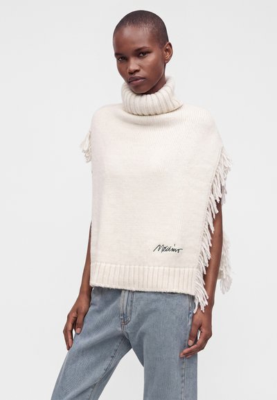 Femme portant un poncho à col roulé crème avec des franges aux manches et le nom de la marque brodé, assorti à un jean bleu clair sur un fond uni.