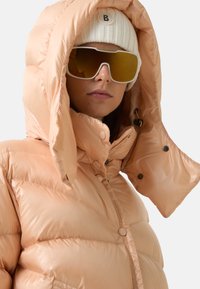 Jasno brzoskwiniowa kurtka puchowa o błyszczącym wykończeniu, z wysokim kołnierzem, oversize'owym kapturem, zatrzaskami oraz diagonalnym pikowaniem.