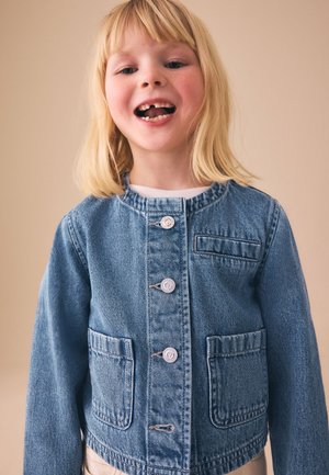 Junges blondes Kind lächelt und zeigt einen fehlenden Vorderzahn, trägt eine blaue Jeansjacke mit Knöpfen und Fronttaschen.