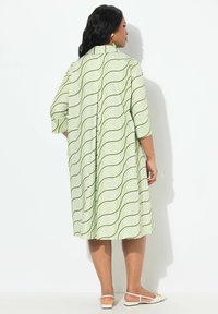 Grünes Kleid mit weißen wellenförmigen Mustern, knielang, dreiviertelärmlig und mit einer geknöpften Rückseite. Getragen mit flachen weißen Sandalen.