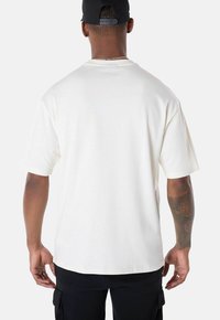 Weißes Kurzarm-T-Shirt mit Rundhalsausschnitt, das eine glatte Textur und einen entspannten Schnitt aufweist, von hinten betrachtet, kombiniert mit schwarzen Hosen.