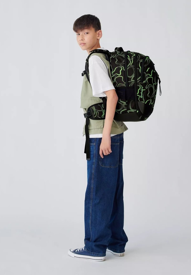 Schwarzer Rucksack mit grünem abstractem Muster, mehreren Taschen und verstellbaren Trägern, getragen über einer grauen Weste und blauen weiten Jeans.