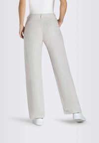 Donna che indossa pantaloni a gamba larga grigi chiari e sneakers bianche, in piedi con le mani nelle tasche, vista di spalle su sfondo bianco.
