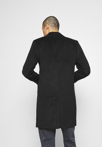 Černý dlouhý kabát s hladkou texturou, se švem na zadní straně a širokými klopami, kombinovaný s tmavými džíny, z pohledu zezadu.