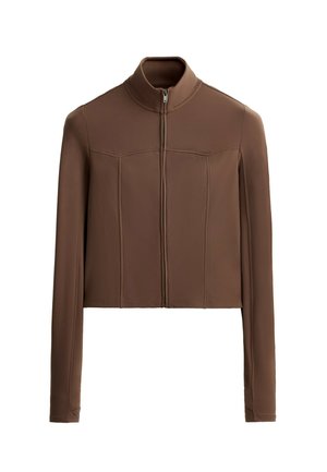 Vrouw staat in een bruine lange mouwen jas met rits en bijpassende fietsbroek, witte sokken en beige sneakers.