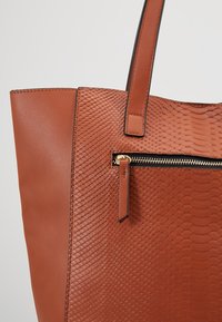 Bruine tote bag met een gestructureerd, slangachtig patroon aan de ene kant, gladde leren afwerking aan de andere kant, en een gouden ritszakaccent.