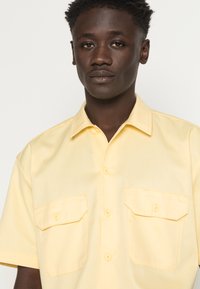 Jovem de pele escura e cabelo encaracolado curto, vestindo uma camisa de botão de manga curta amarela pálida com bolsos frontais.