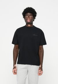 Sort bomulds T-shirt med korte ærmer, prydet med et diskret sort logo på brystet. Modellen har grå sweatpants på. Enkel, afslappet pasform.