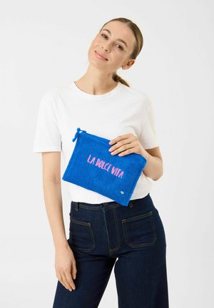Frau in weißem T-Shirt und dunklen Jeans hält ein blaues Täschchen mit pinkem Schriftzug „LA DOLCE VITA“ vor einem einfarbigen weißen Hintergrund.
