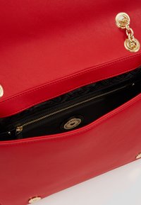 Punainen nahkalaukku kultaisilla yksityiskohdilla, jossa on iso pääosasto ja vetoketjullinen tasku mustalla kuviollisella vuorilla.