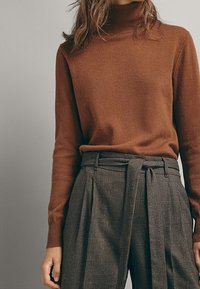 Femme portant un pull à col roulé marron rentré dans un pantalon gris à carreaux taille haute avec une ceinture nouée.