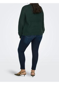 Femme portant un pull en maille vert foncé, un jean skinny bleu foncé et des escarpins à talons bas verts olive, vue de dos.