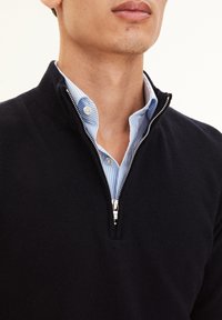 Pull marin avec un col zippé sur une chemise rayée bleu clair. Le pull présente une texture lisse et des accents de poignets côtelés.