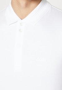 Vit polo-skjorta med ribbad krage, med två klara knappar och en präglad "BOSS"-logotyp på bröstet. Slätt tyg.