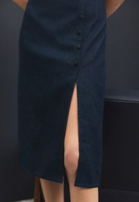 Jupe en denim bleu foncé avec une fente latérale. Présente une fermeture à boutons sur le côté. Tissu lisse, coupe structurée.