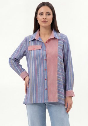 Woman wearing a long-sleeve, button-up shirt with vertical blue and red stripes and light blue jeans, standing with one hand on hip.