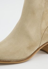 Botín de ante beige con puntera curvada, tacón de madera apilada y detalles de costura visibles junto a una textura suave.