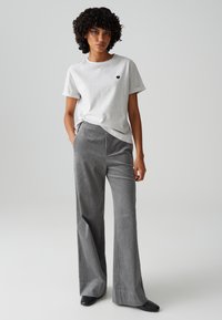 Graues Baumwoll-T-Shirt mit gekrempelten Ärmeln und kleinem schwarzen Herzlogo, kombiniert mit grauen Cordhosen mit weitem Bein und schwarzen High Heels.