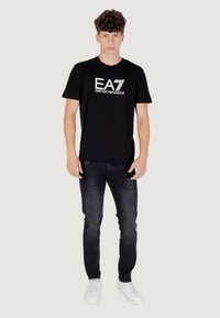 Camiseta de algodón negra con cuello redondo, que presenta un logo blanco de EA7 Emporio Armani. Combinada con jeans oscuros y zapatillas blancas.