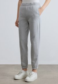 Pelēkas sporta bikses ar elastīgu jostu, sānu kabatām un ribotām aprocēm. Kopā ar baltām sporta kurpēm uz neitrālas grīdas.