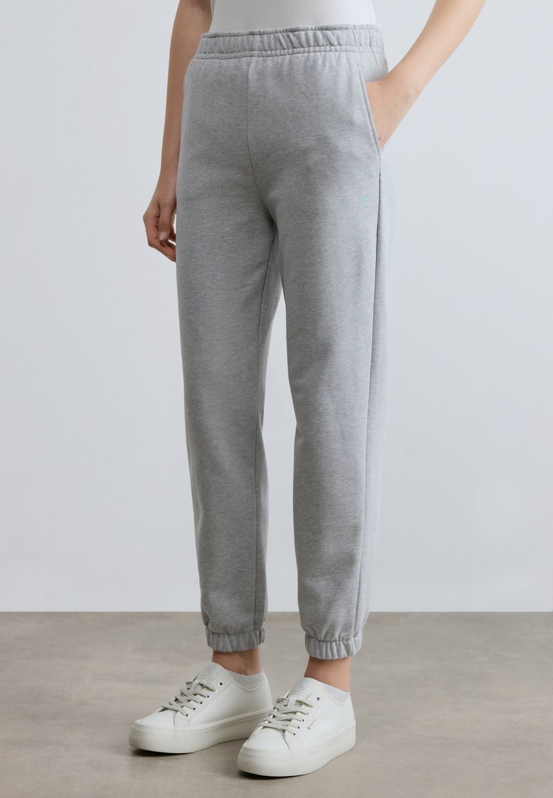 Pelēkas sporta bikses ar elastīgu jostu, sānu kabatām un ribotām aprocēm. Kopā ar baltām sporta kurpēm uz neitrālas grīdas.