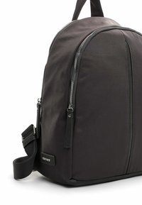 Mochila de tela negra con forma curva, que cuenta con cremalleras plateadas, acentos de cuero y una correa ajustable. Diseño minimalista con una base resistente.