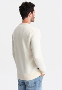 Sweatshirt couleur crème avec des manches longues, des poignets et un ourlet côtelés. Présente un design simple, une texture douce et des accents matériels minimaux.