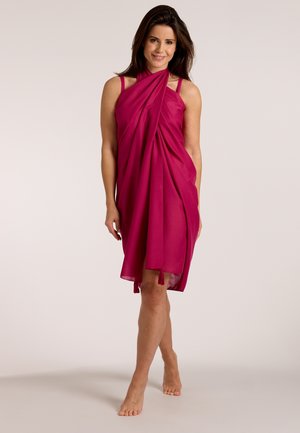 Woman stands barefoot wearing a sleeveless magenta wrap dress with tassels on a plain light background, smiling at the camera.