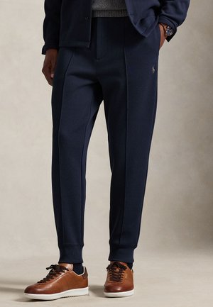 Pantaloni sportivi bleumarin, croiți, cu textură netedă, dotati cu buzunare laterale și un logo subtil, asortați cu pantofi sport maro din piele.