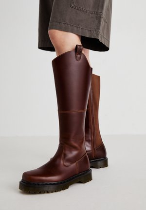 Bottes en cuir marron montant jusqu'aux genoux avec des semelles épaisses noires, portées avec une jupe sombre longueur genou sur un fond uni.