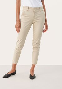 Chino beige slim-fit avec un devant plat, des passants de ceinture et des jambes fuselées, associés à des chaussures plates noires. Tissu en coton lisse, design minimal.