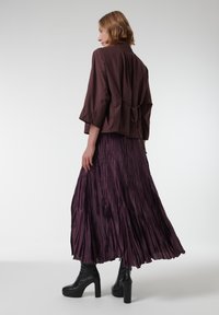 Bruine gecropte jas met een strikdetail aan de achterkant, gecombineerd met een paarse geplooide maxi rok. Zwarte platform enkellaarsjes. Glad textuur overal.