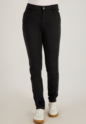 Zwarte nauwsluitende broek met zakken aan de voorkant en knoopsluiting, gedragen met witte sneakers en een wit shirt dat in de broek is gestopt.