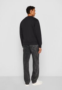 Male model facing away wearing a black sweatshirt, faded black jeans, and white sneakers on a plain gray background.