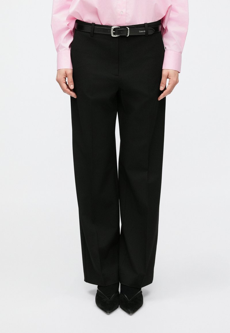 Čierne nohavice s širokými nohavicami a hladkým textúrou, doplnené o opaskové slučky, v kombinácii s ružovou košeľou s dlhým rukávom. Vidieť sú čierne topánky s ostrou špičkou.