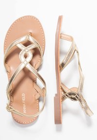 Flade sandaler i tan-farve med metallisk guld crisscross stropper, ankelrem med spænde og "Anna Field" logo på fodsengen, vist fra top- og sidevinkler.