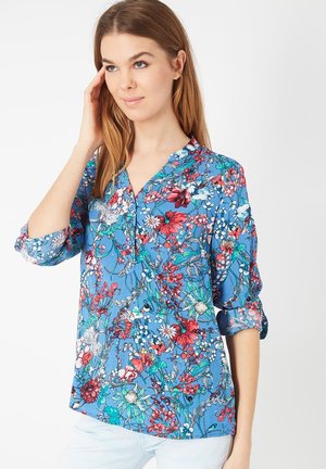 Blusa floral azul con escote en V, mangas tres cuartos, que presenta patrones vibrantes de flores rojas, rosas y blancas sobre un fondo azul claro.