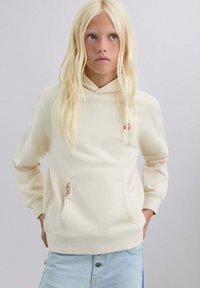 Persona rubia con cabello largo, vistiendo una sudadera color crema con bolsillo frontal y jeans azul claro, de pie con las manos en los bolsillos.