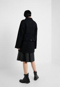 Abrigo negro de mezcla de lana con cuello, gran bolsillo trasero de parche y detalles de botones, combinado con una falda de cuero plisada negra y botas de combate negras.