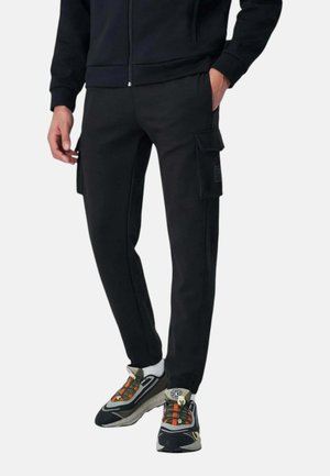 Uomo che indossa pantaloni cargo neri con tasche a patta, una giacca nera, calzini bianchi e sneakers multicolore, in piedi davanti a uno sfondo semplice.