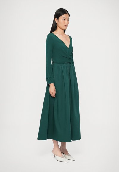 Φόρεμα ελιάς πράσινο μέχ�ρι το πάτωμα με μακριά μανίκια, v-λαιμόκοψη και φούσκωμα στη μέση. Συνδυασμένο με λευκά παπούτσια με χαμηλό τακούνι. Λείο ύφασμα.