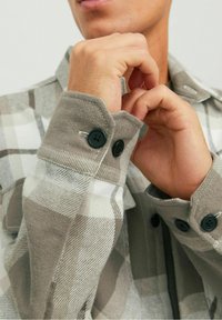 Camicia di flanella a quadri grigia e bianca con una texture morbida, caratterizzata da bottoni neri e un colletto classico. Mani che aggiustano i polsini.