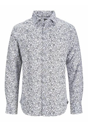 Mann trägt ein weißes Langarmhemd mit Blumenmuster, schwarze Hose und weiße Turnschuhe und steht vor einem einfarbigen Hintergrund.