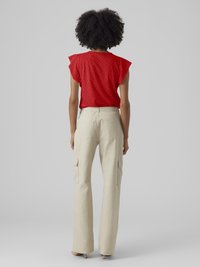 Γυναίκα με κατσαρά μαλλιά στέκεται με την πλάτη, φορώντας μια κόκκινη μπλούζα με τρυπητά σχέδια, μπεζ παντελόνι cargo και μπεζ τακούνια πάνω σε απλό γκρι φόντο.